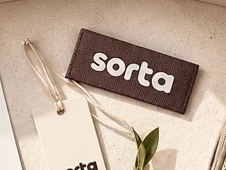 A woven Sorta label and a paper swing tag beside an olive sprig.